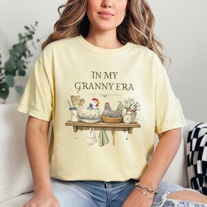May include: A light yellow t-shirt with the phrase "IN MY GRANNY ERA" printed on the front. The graphic features a shelf with chickens, kitchen utensils, and flowers. The shirt is a relaxed fit.