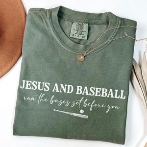 May include: Olive green t-shirt with the text "JESUS AND BASEBALL run the bases set before you" in white. A baseball bat and ball graphic is below the text. A gold necklace with a pendant is on the shirt.