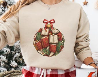 May include: Beige sweatshirt featuring a Christmas ornament design made of books. The ornament is composed of red and green books, some open, with a red bow at the top. The design is festive and book-themed.