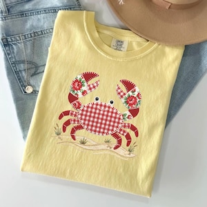 Op de afbeelding: Geel Comfort Colors T-shirt met een krab applicatie. De krab heeft een rood-wit geruit lichaam en rode klauwen met bloemaccenten. Het shirt wordt getoond met een blauwe spijkerbroek en een bruine hoed.