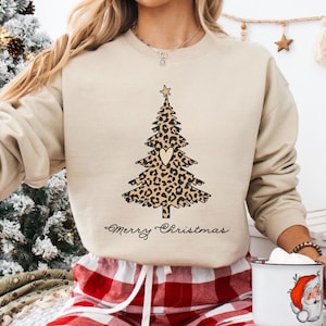 Può includere: Una felpa beige con un design ad albero di Natale con stampa leopardata e un ritaglio a forma di cuore. Le parole "Merry Christmas" sono scritte in corsivo sotto l'albero. La felpa è abbinata a pantaloni da pigiama a quadri rossi e bianchi.