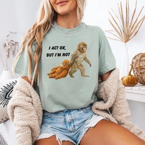 May include: A light green t-shirt with the text "I ACT OK, BUT I'M NOT" and a graphic of two monkeys. One monkey is standing, and the other is lying down. The shirt is a relaxed fit.