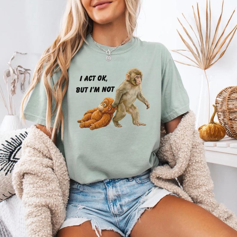 May include: A light green t-shirt with the text "I ACT OK, BUT I'M NOT" and a graphic of two monkeys. One monkey is standing, and the other is lying down. The shirt is a relaxed fit.