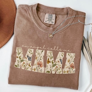 May include: A brown Comfort Colors t-shirt with the text "an angel calls me MAMA". The word "MAMA" is filled with a floral and butterfly design. A silver necklace with a pendant is on the shirt.