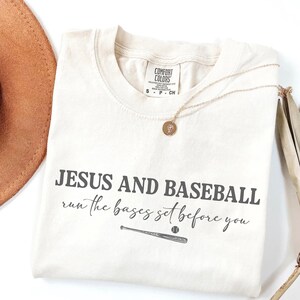 May include: Cream-colored t-shirt with the text "JESUS AND BASEBALL run the bases set before you" in dark gray. A baseball bat and ball graphic is below the text. A gold necklace with a pendant is on the shirt.
