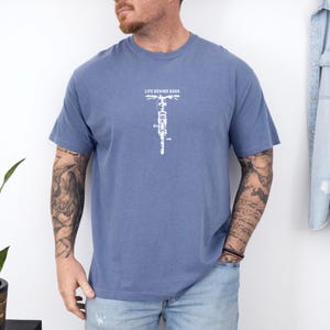 May include: A blue short-sleeve t-shirt with a white graphic of a bicycle and the text "LIFE BEHIND BARS" above it. The shirt is made of a soft material.