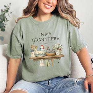 May include: Sage green t-shirt with the text "IN MY GRANNY ERA". The shirt features a shelf graphic with chickens, kitchen utensils, and flowers. The design includes a blue and white ceramic bowl and a woven basket.