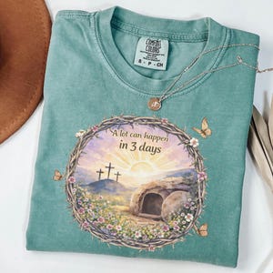 Può includere: Una maglietta Comfort Colors turchese con la scritta "A lot can happen in 3 days" sopra una grafica di un'alba, delle croci e una tomba. La maglietta ha un girocollo e maniche corte. Una collana d'argento con un ciondolo è drappeggiata sulla maglietta.
