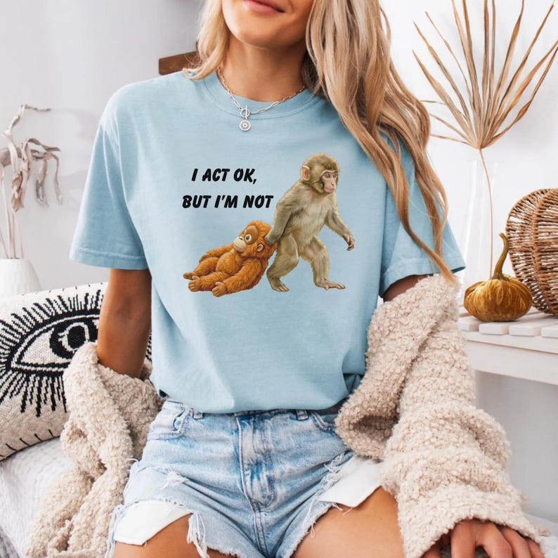 May include: Light blue t-shirt with the text "I ACT OK, BUT I'M NOT" and a graphic of two monkeys. One monkey is standing, and the other is lying down. The shirt is made of a soft material.