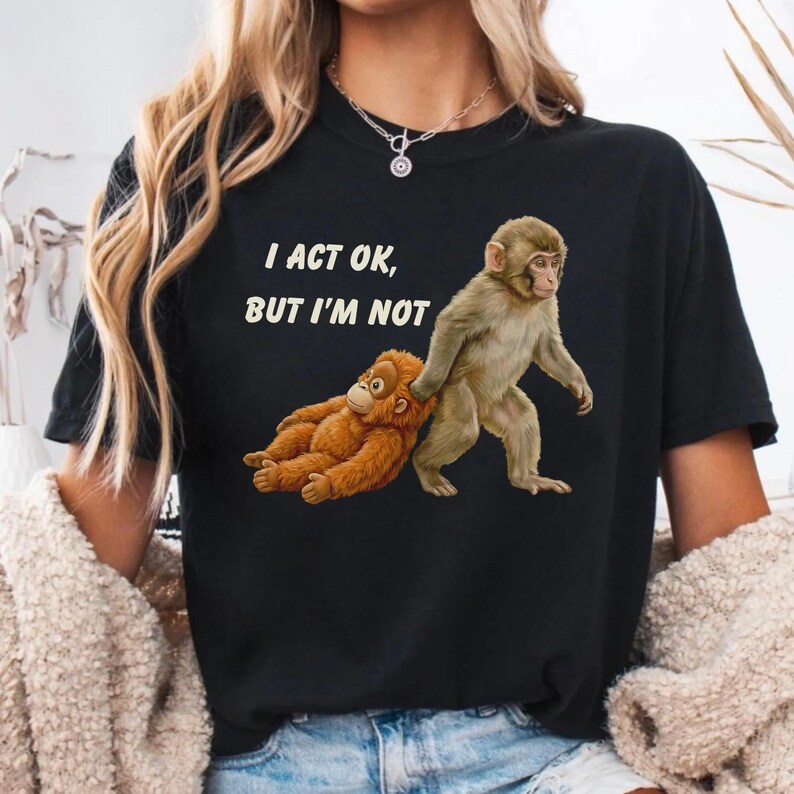 May include: Black t-shirt with the text "I ACT OK, BUT I'M NOT" in white. The shirt features a graphic of two monkeys, one lying down and the other standing. The lying monkey is brown, and the standing monkey is tan.