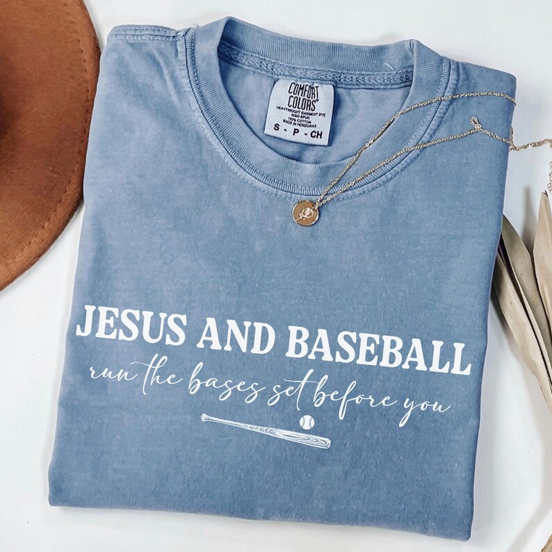 May include: A blue Comfort Colors t-shirt with the text "JESUS AND BASEBALL run the bases set before you" in white. A baseball bat and ball graphic is below the text. A gold necklace with a pendant is on the shirt.