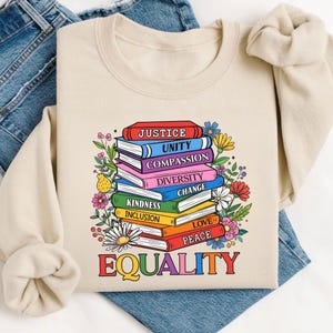 May include: Beige sweatshirt with a colorful graphic of stacked books. Each book displays a word like "Justice," "Unity," and "Equality." Floral accents surround the books, and the word "EQUALITY" is at the bottom in rainbow colors.