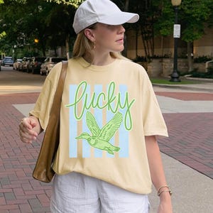 May include: A light yellow t-shirt with the word "lucky" in green script and a green duck graphic. The shirt has blue vertical stripes behind the duck. The person is wearing a white baseball cap and white shorts.