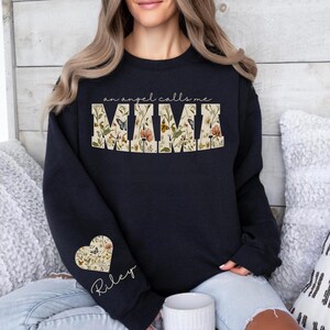 Pode incluir: Um moletom azul marinho com o texto "an angel calls me MIMA" em um design floral. Um design floral em forma de cora&ccedil;&atilde;o com o nome "Riley" est&aacute; na manga esquerda. &Eacute; um estilo casual e confort&aacute;vel.
