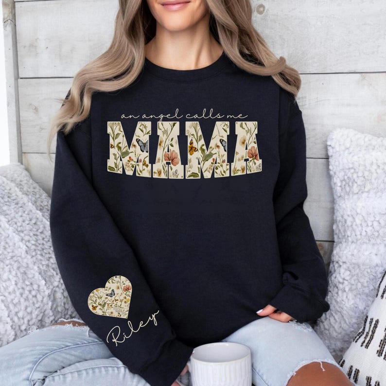 Pode incluir: Um moletom azul marinho com o texto "an angel calls me MIMA" em um design floral. Um design floral em forma de cora&ccedil;&atilde;o com o nome "Riley" est&aacute; na manga esquerda. &Eacute; um estilo casual e confort&aacute;vel.