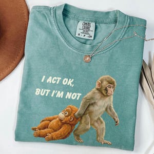 May include: A teal Comfort Colors t-shirt featuring a graphic of two monkeys, one walking and the other being dragged. The text "I ACT OK, BUT I'M NOT" is printed above the monkeys. A silver necklace with a pendant is also visible.