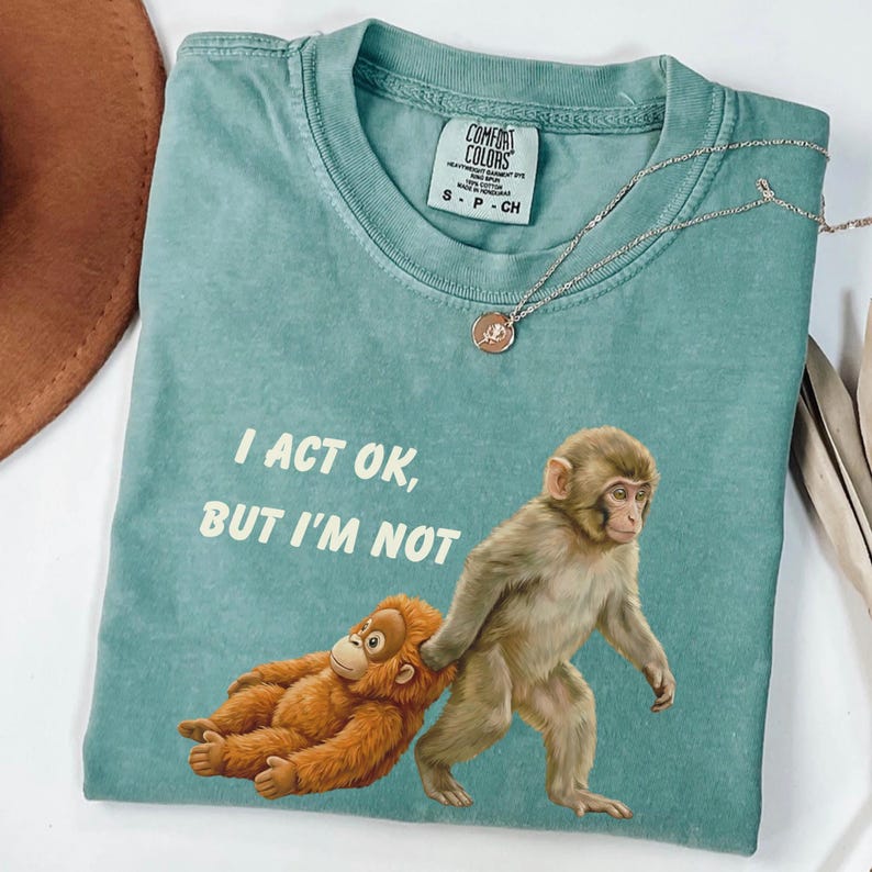 May include: A teal Comfort Colors t-shirt featuring a graphic of two monkeys, one walking and the other being dragged. The text "I ACT OK, BUT I'M NOT" is printed above the monkeys. A silver necklace with a pendant is also visible.