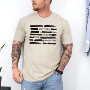Può includere: T-shirt beige con la scritta "everything is fine trust your government" in nero, con lettering a pennello. La maglietta è di colore neutro e ha maniche corte. Il disegno è centrato sul davanti.