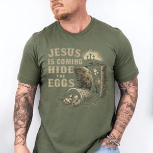 May include: Olive green t-shirt with the text "JESUS IS COMING HIDE THE EGGS" in a vintage style font. The graphic includes an illustration of a tomb, three crosses, and a basket of eggs.
