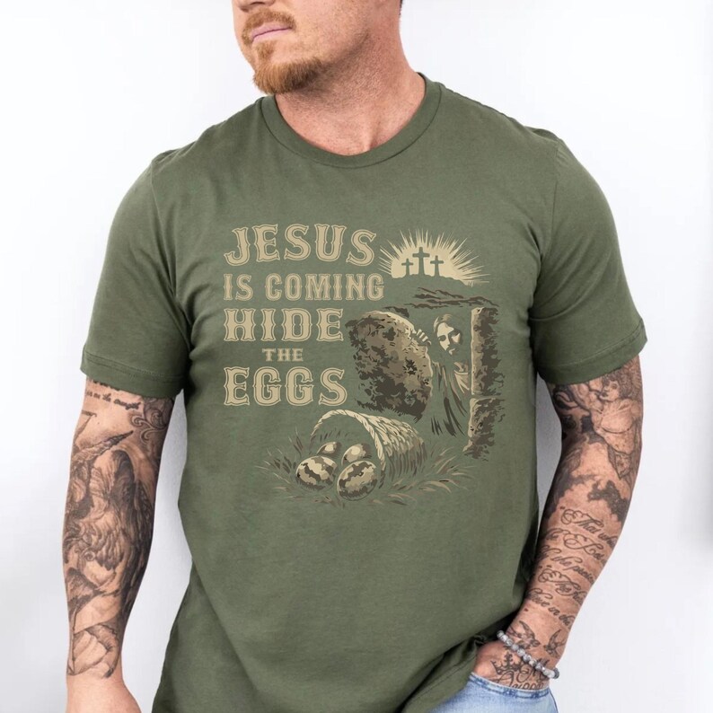 May include: Olive green t-shirt with the text "JESUS IS COMING HIDE THE EGGS" in a vintage style font. The graphic includes an illustration of a tomb, three crosses, and a basket of eggs.