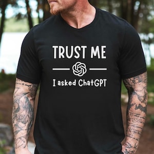 May include: Black crew neck t-shirt with the white text "TRUST ME" above a graphic and the text "I asked ChatGPT". The shirt is made of a soft material.