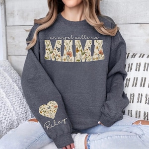 Pode incluir: Camisola cinza escuro com o texto "an angel calls me MAMA" em um design floral. Um design floral em forma de cora&ccedil;&atilde;o com o nome "Riley" est&aacute; na manga esquerda. A camisola &eacute; um estilo confort&aacute;vel e casual.