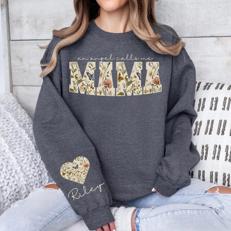 Pode incluir: Camisola cinza escuro com o texto "an angel calls me MAMA" em um design floral. Um design floral em forma de cora&ccedil;&atilde;o com o nome "Riley" est&aacute; na manga esquerda. A camisola &eacute; um estilo confort&aacute;vel e casual.
