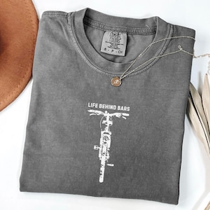 May include: A gray Comfort Colors t-shirt with the text "LIFE BEHIND BARS" and a white bicycle graphic. A gold-colored necklace with a pendant is draped across the shirt. A brown hat is visible in the upper left corner.