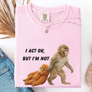 May include: A light pink t-shirt with the text "I ACT OK, BUT I'M NOT" and a graphic of two monkeys. One monkey is walking, and the other is lying down. A silver necklace with a pendant is on the shirt.