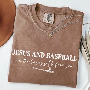 May include: A brown Comfort Colors t-shirt with the text "JESUS AND BASEBALL run the bases set before you" in white. A baseball bat and ball graphic is below the text. A silver necklace with a pendant is on the shirt.