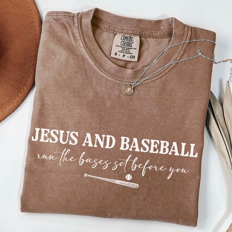 May include: A brown Comfort Colors t-shirt with the text "JESUS AND BASEBALL run the bases set before you" in white. A baseball bat and ball graphic is below the text. A silver necklace with a pendant is on the shirt.