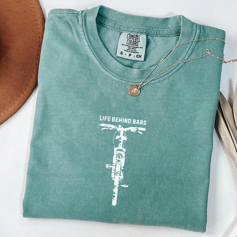 May include: A seafoam green Comfort Colors t-shirt with the text "LIFE BEHIND BARS" and a white bicycle graphic. A gold necklace and a brown hat are also visible. The shirt is size small.