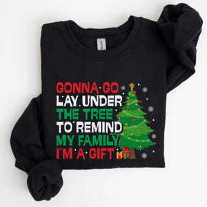 May include: Black long-sleeve sweatshirt with a Christmas-themed graphic. The graphic includes the text "Gonna go lay under the tree to remind my family I'm a gift" in red and green, along with a decorated Christmas tree and gifts.