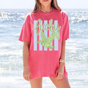May include: A coral-colored t-shirt with a graphic design featuring the word "Lucky" in green script above a green duck in flight. The word and duck are set against a light blue striped background.