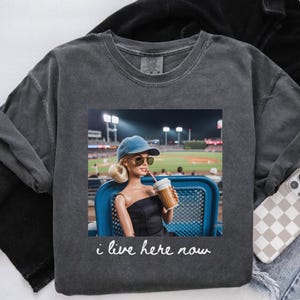 Pu&ograve; includere: Felpa grigio scuro con una grafica di una bambola a una partita di baseball, che tiene una tazza di caff&egrave;. La bambola indossa occhiali da sole e un berretto blu. Il testo "i live here now" &egrave; sotto.