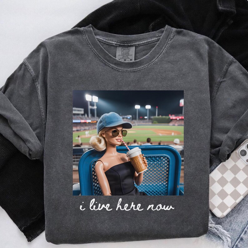 Pu&ograve; includere: Felpa grigio scuro con una grafica di una bambola a una partita di baseball, che tiene una tazza di caff&egrave;. La bambola indossa occhiali da sole e un berretto blu. Il testo "i live here now" &egrave; sotto.