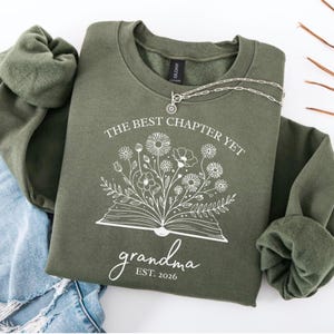 May include: An olive green sweatshirt featuring the text "THE BEST CHAPTER YET" and "grandma EST. 2026" in white. The front displays a floral design above an open book illustration. A silver necklace is draped across the sweatshirt.