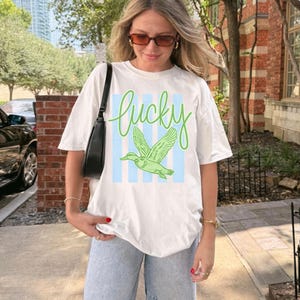May include: White t-shirt with the word "lucky" in green script above a green duck graphic. The duck is set against a backdrop of light blue vertical stripes. The shirt is worn with light blue jeans.