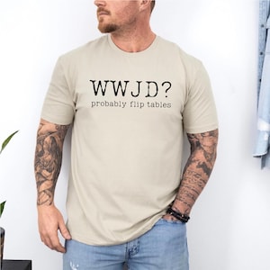 Könnte beinhalten: Beiges T-Shirt mit dem schwarzen Text "WWJD?" über "probably flip tables". Das T-Shirt hat kurze Ärmel und scheint aus einem weichen Material zu sein. Die Person, die das T-Shirt trägt, hat Tätowierungen auf den Armen.