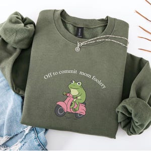 May include: An olive green sweatshirt featuring the text "Off to commit mom foolery" and a cartoon frog on a pink scooter. A silver necklace with a circular pendant is draped on the sweatshirt.