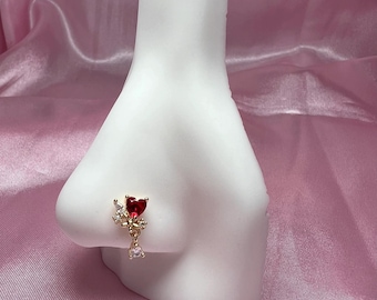 May include: A gold-colored nose ring featuring a red heart-shaped gemstone and small clear crystals. The nose ring is attached to a white nose form, set against a pink satin background. The jewelry is designed to be worn on the nose.