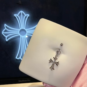 Könnte beinhalten: Ein silberner Bauchnabelpiercing mit einem Anhänger in Kreuzform, verziert mit kleinen, klaren Steinen. Das Kreuzdesign spiegelt sich in einer neonblauen Kontur auf dunklem Hintergrund wider. Der Schmuck wird auf einer weißen Oberfläche präsentiert.