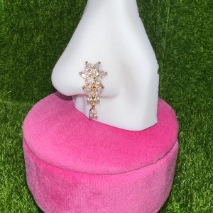 May include: A white nose ring display stand with a clear crystal nose ring with a gold setting. The stand is pink velvet and has a gold base.