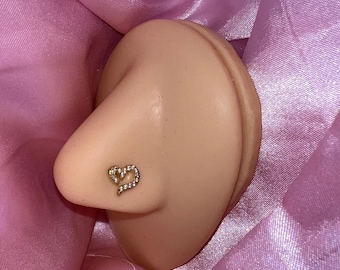 May include: A gold-colored nose stud in the shape of a heart, encrusted with small, clear gemstones. The stud is set on a light-toned prosthetic nose, resting on a bed of pink satin fabric.