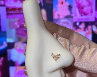 May include: A gold nose stud in the shape of a heart, adorned with clear and light pink gemstones. The stud is set on a white nose form, showcasing the jewelry's design. The background features a collage of pink and purple images.
