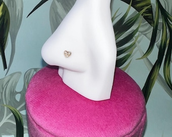 May include: A white nose ring display stand with a small, sparkly heart-shaped nose ring. The stand is sitting on a pink velvet base with silver feet.