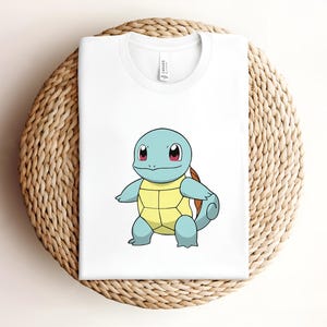 May include: White t-shirt with a cartoon image of Squirtle, a blue turtle Pokemon with a yellow shell, red eyes, and a brown shell.