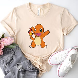 May include: A beige t-shirt with a cartoon image of a fire-type Pokemon, Charmander, with a flame on its tail. The Pokemon is orange and yellow with black eyes and a wide smile.