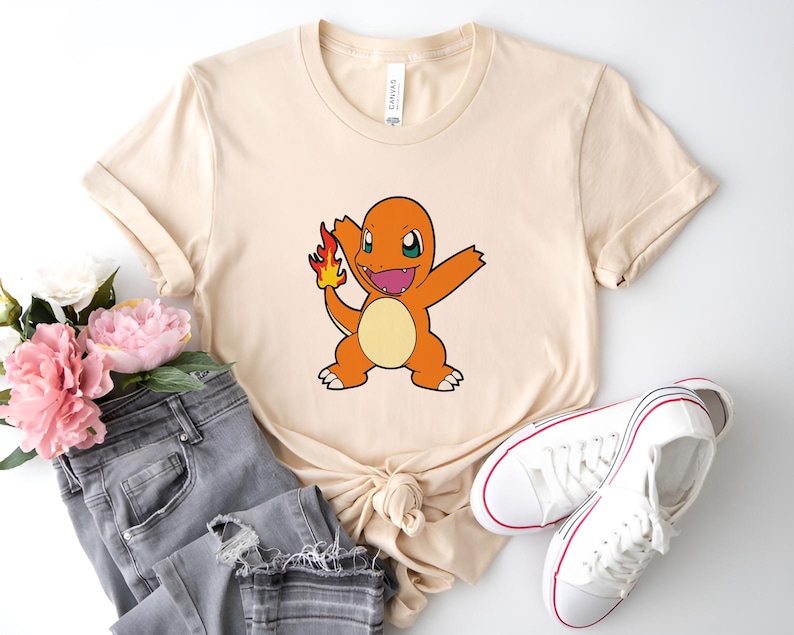 May include: A beige t-shirt with a cartoon image of a fire-type Pokemon, Charmander, with a flame on its tail. The Pokemon is orange and yellow with black eyes and a wide smile.