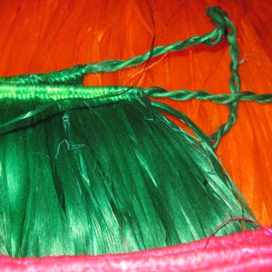 May include: A green grass-like skirt featuring a braided waistband and long, thin, fibrous strands. The skirt is positioned against a bright orange background, with a portion of a pink braided trim visible along the bottom edge of the frame.
